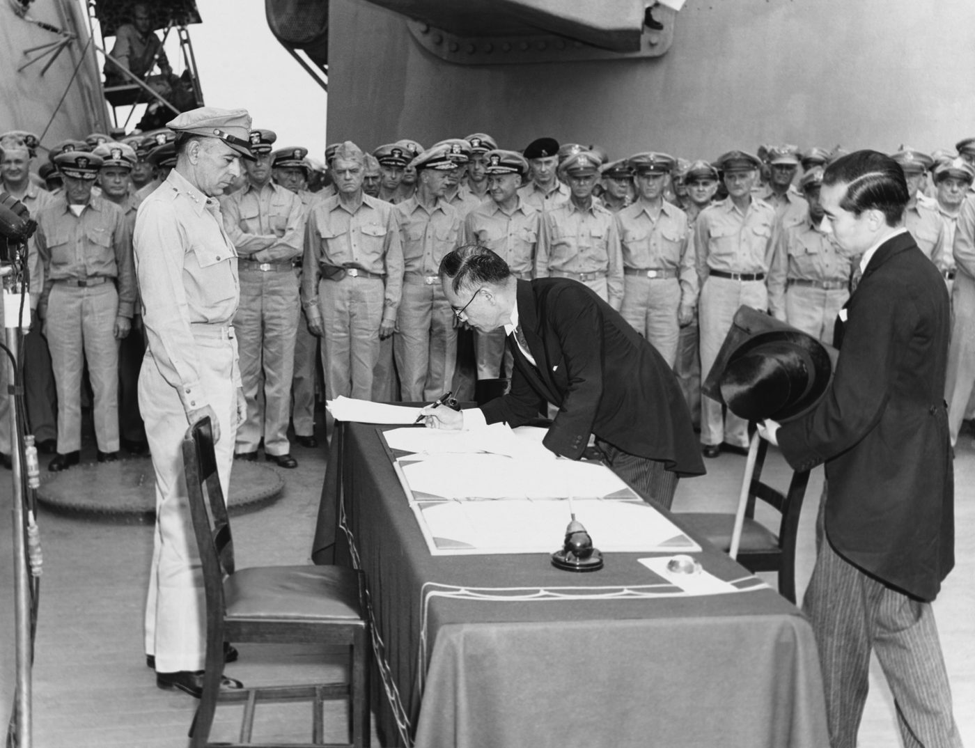 軍艦の上で、大勢の軍服の人々が見守る中、黒い服の男性が机にかがんで書類に署名し、その横でぼうしを持った男性が立っている白黒の写真