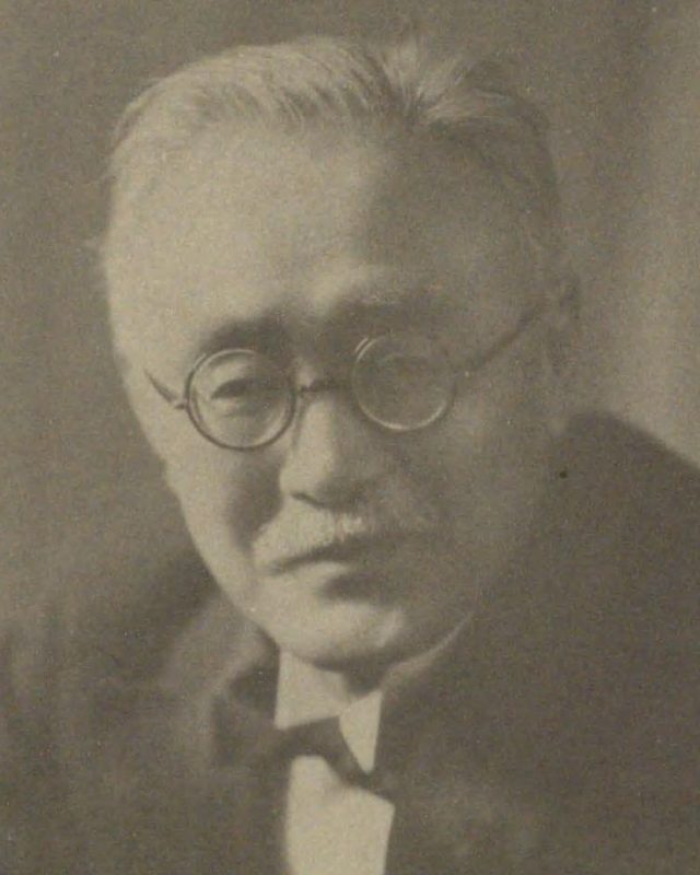 丸いめがねをかけ、白い髪で口ひげのある、洋服の男性の顔