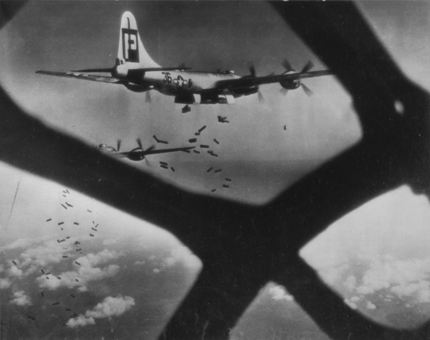 飛行機の窓のわくごしに、尾翼に大きな文字のある4つのプロペラの爆撃機がとび、その下に小さな爆弾がたくさん落ちていく白黒の写真