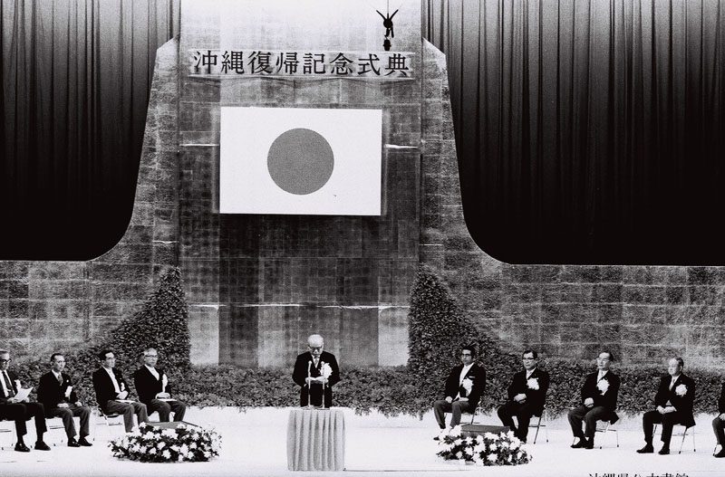 舞台の上に「沖縄復帰記念式典」と書かれた幕と日の丸がかかげられ、まん中で白い髪の男性がことばを述べ、左右にスーツの人々がすわっている白黒の写真