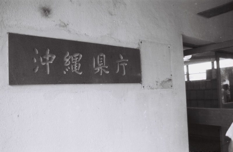 白いかべに取りつけられた、「沖縄県庁」と書かれた横長の看板の白黒の写真