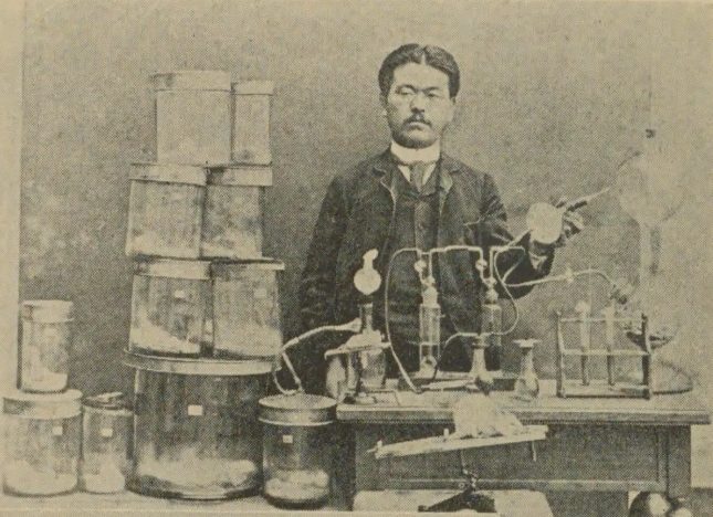 洋服を着た男性が、ガラスの器具や容器がならんだ机の前に立っている古い写真