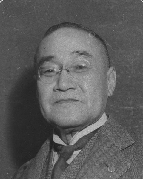 めがねをかけ、スーツにネクタイの年配の男性