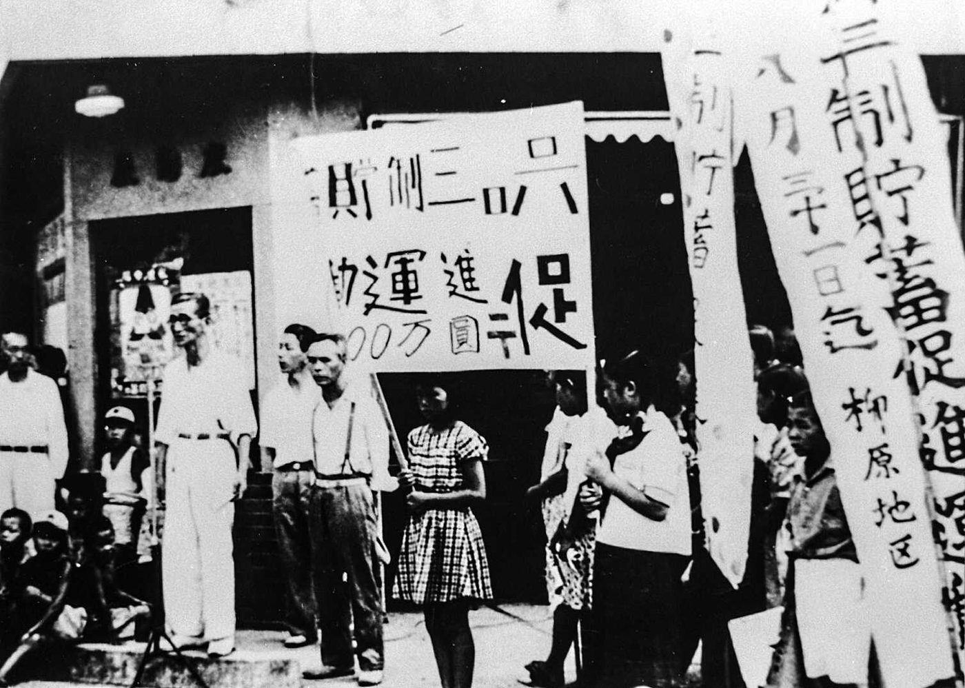 「六三制」「促進運動」と書かれた大きな紙や、「貯蓄促進」と書かれたのぼりを持った人々が、店の前に立っている白黒の写真