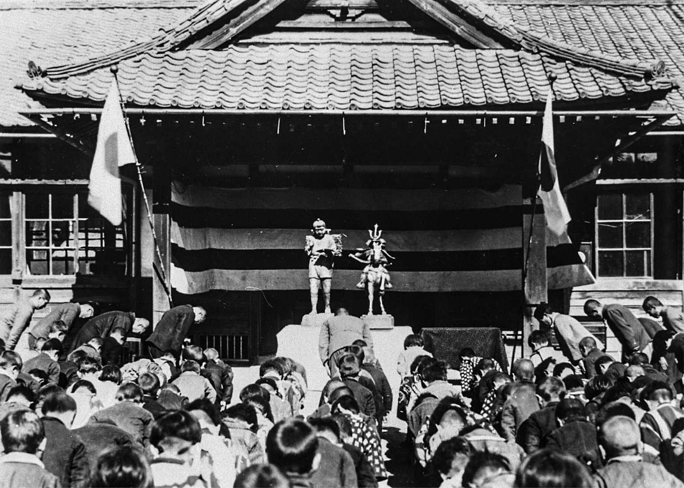 日の丸の旗が立てられた校舎の前に、まきを背負った少年の像と、馬に乗った武将の像が置かれ、大勢の子どもと大人が頭を下げている白黒の写真